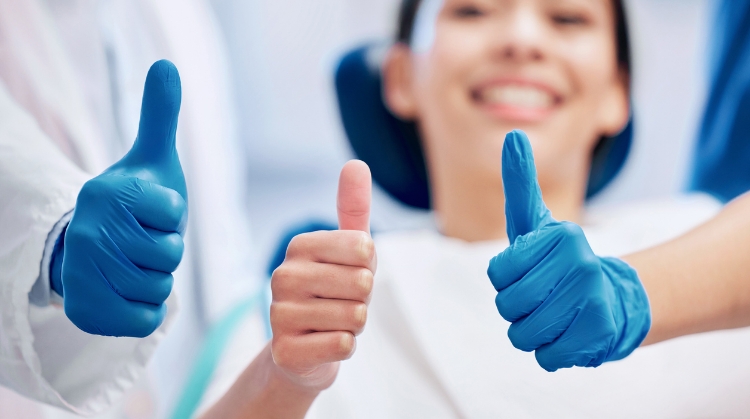 Smiling dental patient giving a thumbs up with two dental professionals also giving thumbs up, wearing blue gloves.