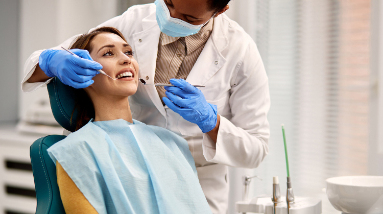 Dentist examining a patient's teeth in a dental office. The patient is seated in a dental chair, smiling, while the dentist uses a dental mirror and explorer for the examination.